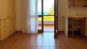 Una habitación con puerta corrediza de vidrio que da a un balcón, un escritorio de madera con una silla, y un pequeño estante con adornos.