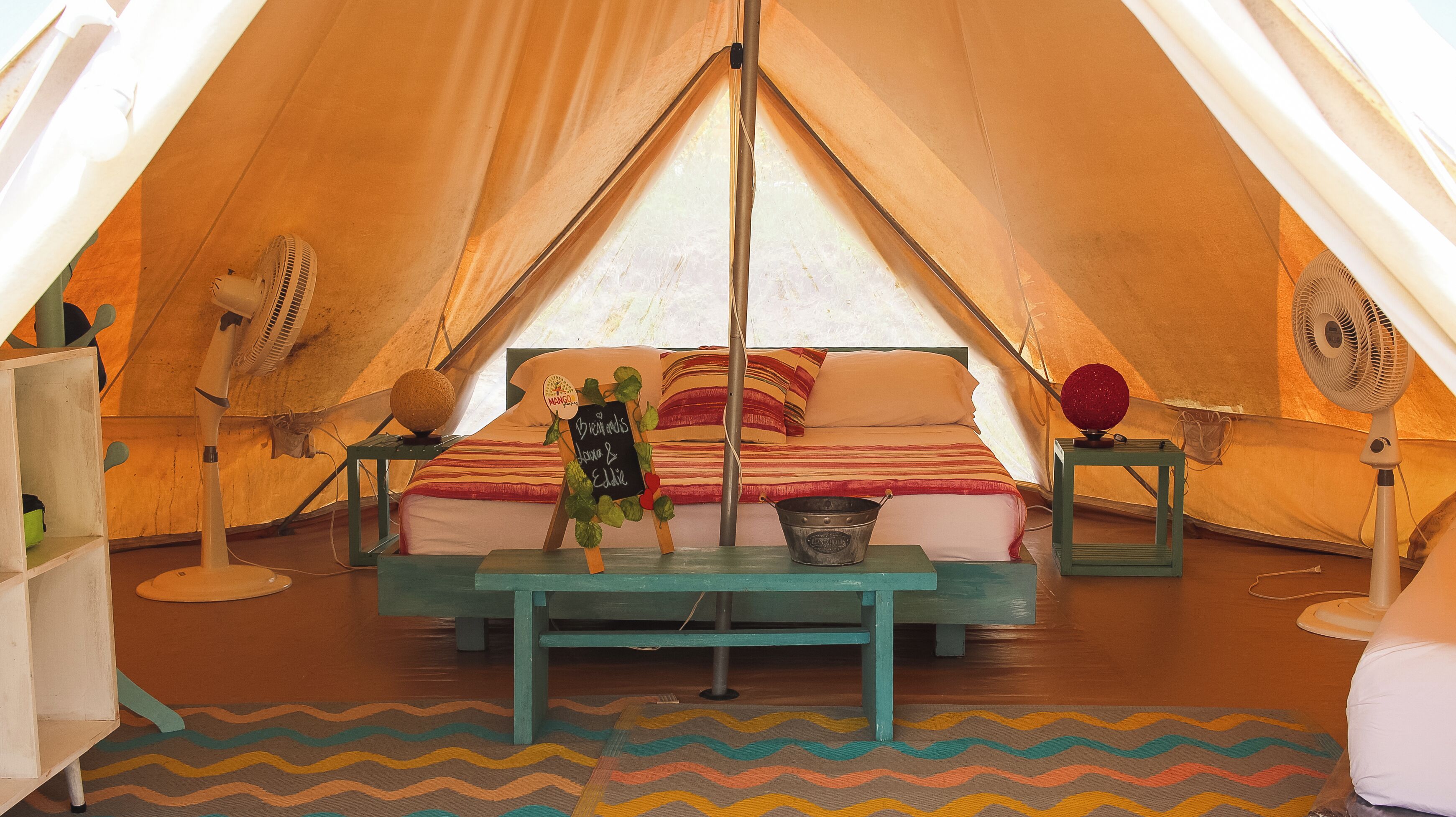 A cozy tent with a bed, two fans, a bucket, and a chalkboard sign.