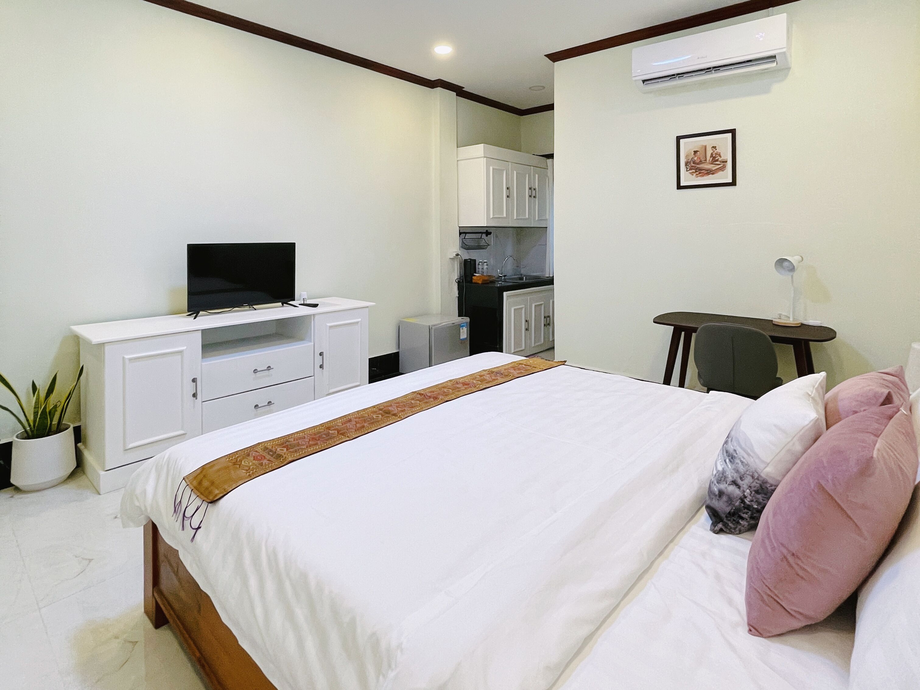 A hotel room with a bed, a TV on a white cabinet, a small kitchenette, and a desk with a chair.