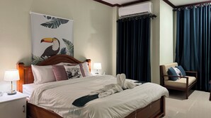 A hotel room with a wooden bed, a nightstand with a lamp, a chair, a large poster on the wall, and a window with dark curtains. - Vanida Hotel (Vientiane)