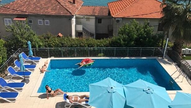 Ein Swimmingpool mit blauen Liegestühlen und Sonnenschirmen, eine Person schwimmt, und im Hintergrund Meer und Gebäude.