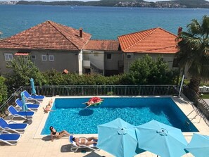 A swimming pool with blue chairs and umbrellas, a person swimming, and a view of the sea and buildings in the background. - Hotel Bavaria (Seget Donji)