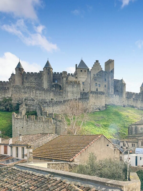 A historic stone castle with multiple towers and a fortified wall, surrounded by a hilly landscape.