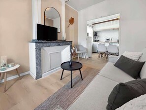 A modern living room with a sofa, a small round table, a flat-screen TV, and a kitchen area with chairs and a dining table.