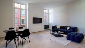 A modern living room with a dining table, black chairs, a blue sofa, a TV mounted on the wall, and large windows.