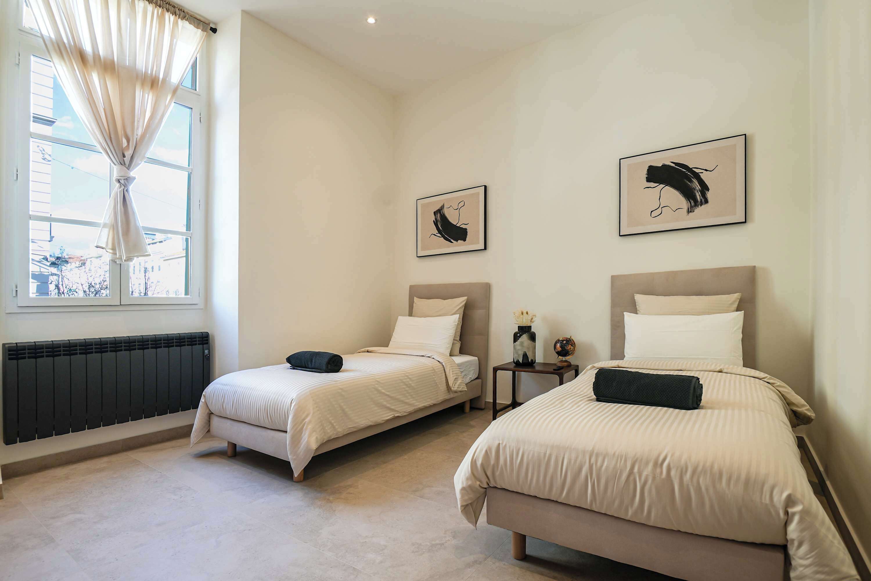 A bedroom with two beds, a radiator, a window with curtains, and two framed artworks on the wall.