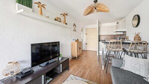 A modern living room with a flat-screen TV, a dark entertainment unit, and a dining area with a wooden table and wicker chairs. - Le Cap Vert - 4 pers, terrasse & piscine (Mandelieu-la-Napoule)