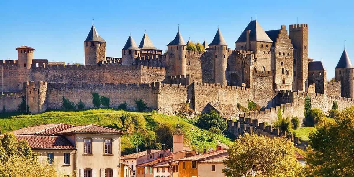 Un castillo histórico con múltiples torres y una muralla fortificada, rodeado de vegetación exuberante y edificios residenciales.