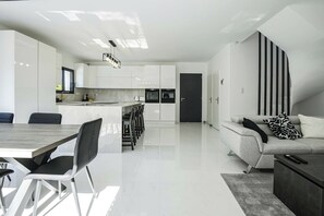 Une cuisine moderne avec des meubles blancs, un coin repas avec une table et des chaises en bois, et un salon avec un canapé et une télécommande.