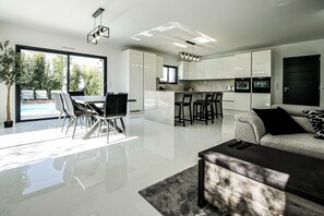 A modern living room with a sofa, coffee table, and a dining area with chairs. There is a kitchen with white cabinets and a large window overlooking a pool.