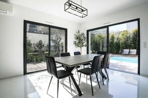 A modern dining area with a large table and chairs, large windows, and a view of a swimming pool. - LX133 Villa jardin, piscine privée et calme absolu (Villeneuve-Loubet)