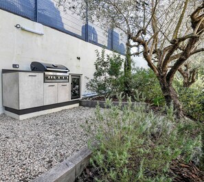 An outdoor barbecue area with a grill and storage cabinets, surrounded by gravel and plants.
