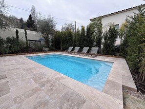 A rectangular swimming pool with a tiled deck, surrounded by trees and a building.