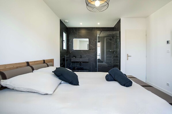A modern hotel room with a large bed, two pillows, a headboard, a bedside table, a mirror, a bathroom with a glass shower enclosure, and a door.