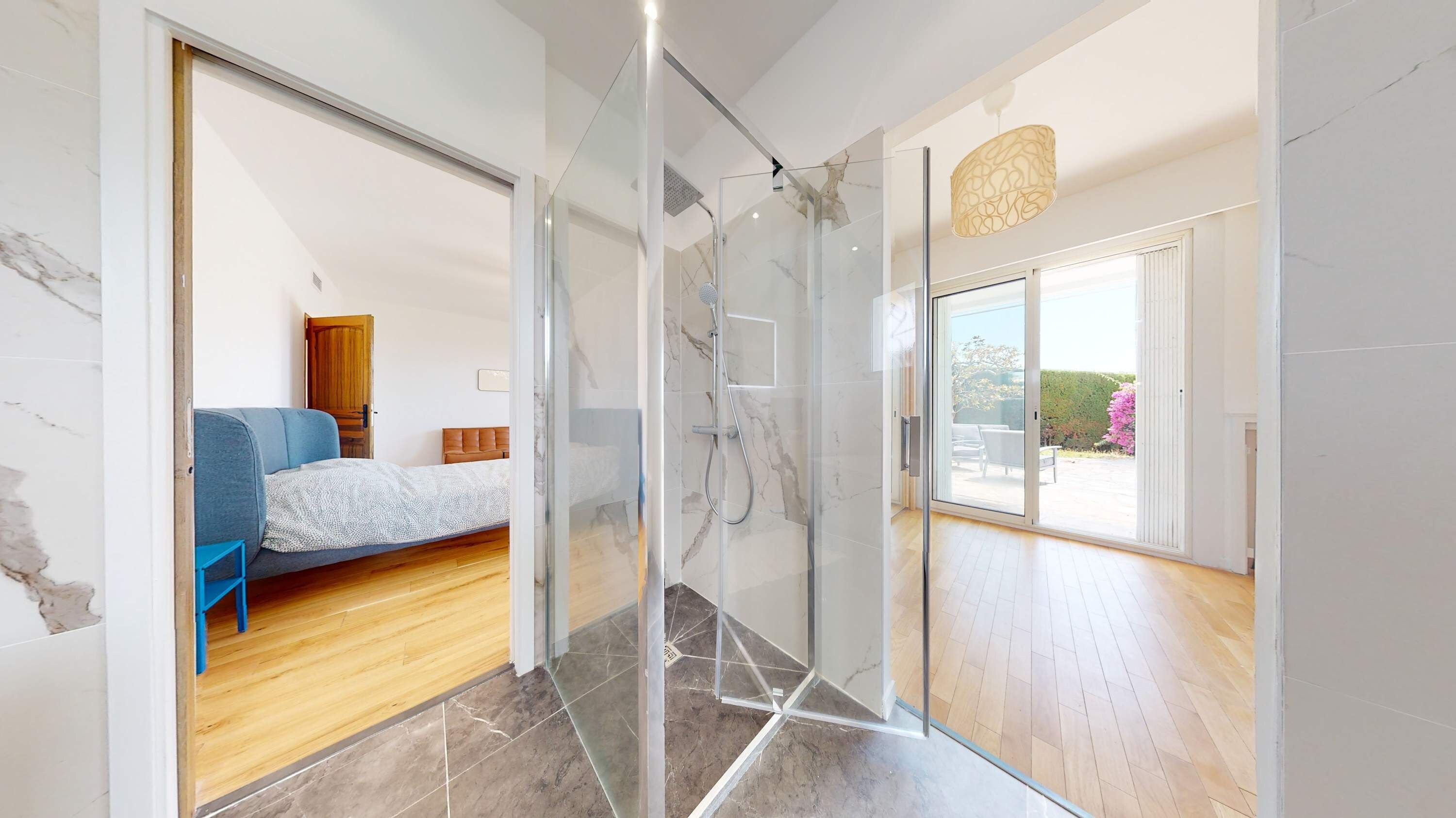 Un baño moderno con cabina de ducha de vidrio, suelo de madera y vistas a un dormitorio con cama y sofá azul.