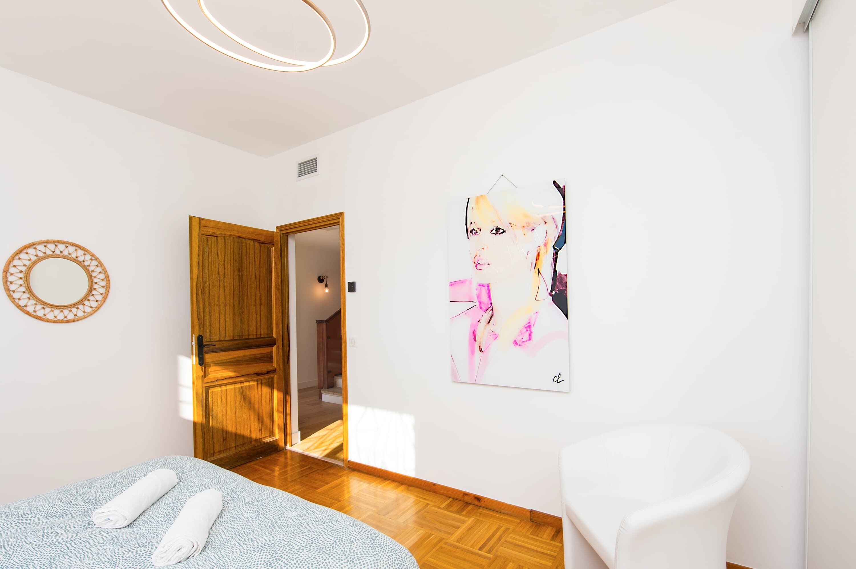 Une pièce avec une porte en bois, un miroir, un tableau, une chaise blanche et un lit avec des serviettes.