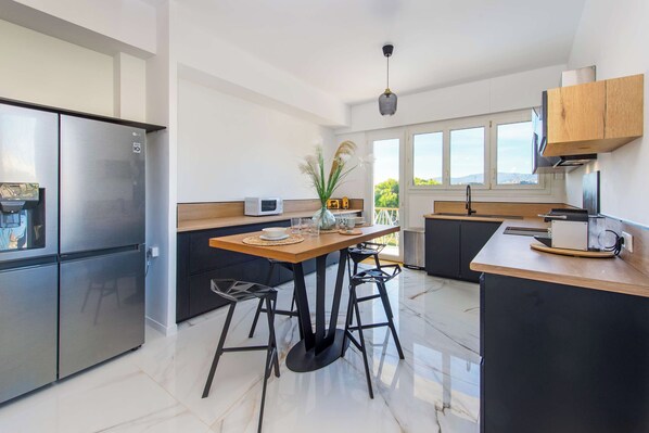 A modern kitchen with a stainless steel refrigerator, a wooden dining table with black stools, and a view of the outdoors through large windows.