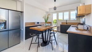 A modern kitchen with a stainless steel refrigerator, a wooden dining table with black stools, and a view of the outdoors through large windows.