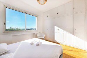 A bedroom with a large window, a bed with rolled towels, a chair, and a wardrobe.