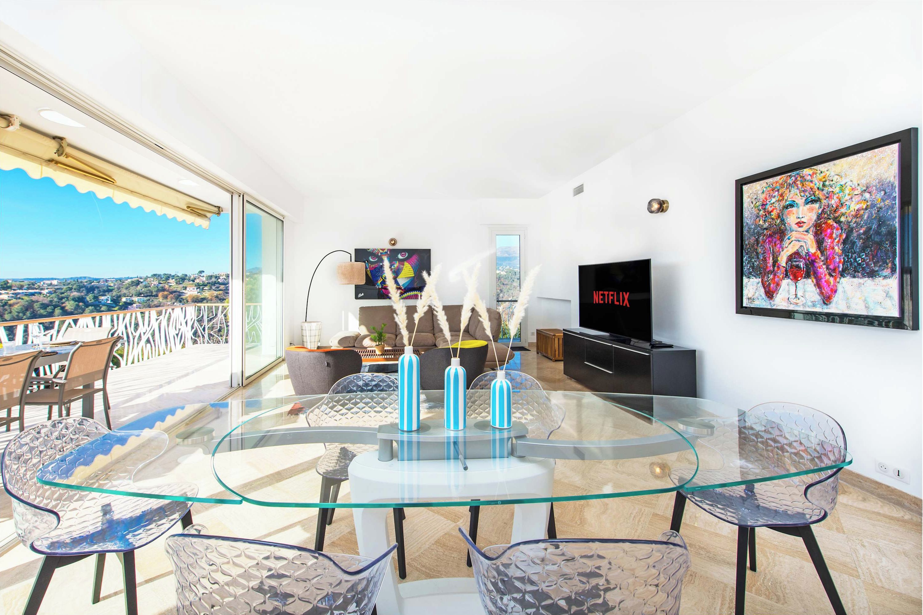 A modern living room with a glass table, clear chairs, a TV, and artwork on the wall.