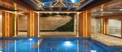 An indoor swimming pool with a geometric ceiling design, a visible wall with 'BENETTONIC ZONE' text, and a clear pool area.