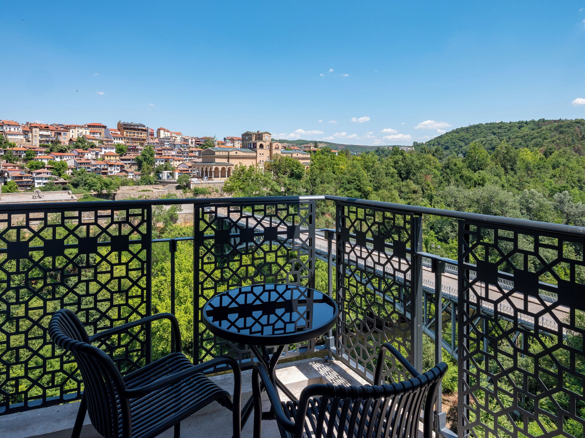 A balcony with a table and chairs, overlooking a town with buildings and a bridge.