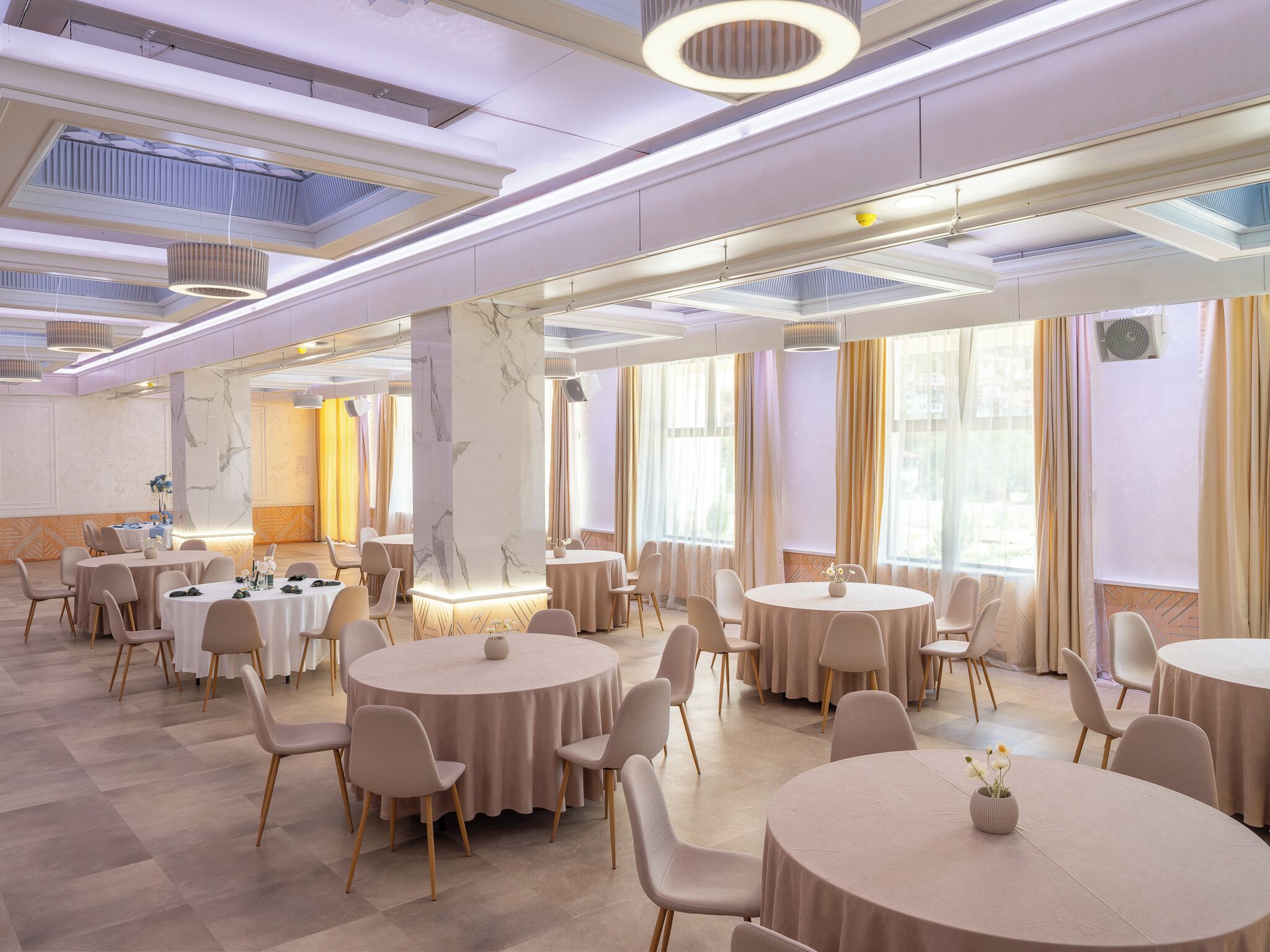Une salle de banquet spacieuse, dotée de tables et de chaises rondes, de grandes fenêtres et d’un haut plafond agrémenté de lucarnes.