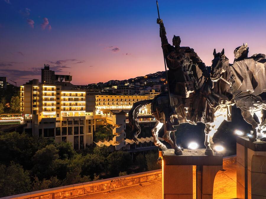 Una estatua de una figura a caballo con una espada levantada, frente a edificios iluminados y un cielo al anochecer.
