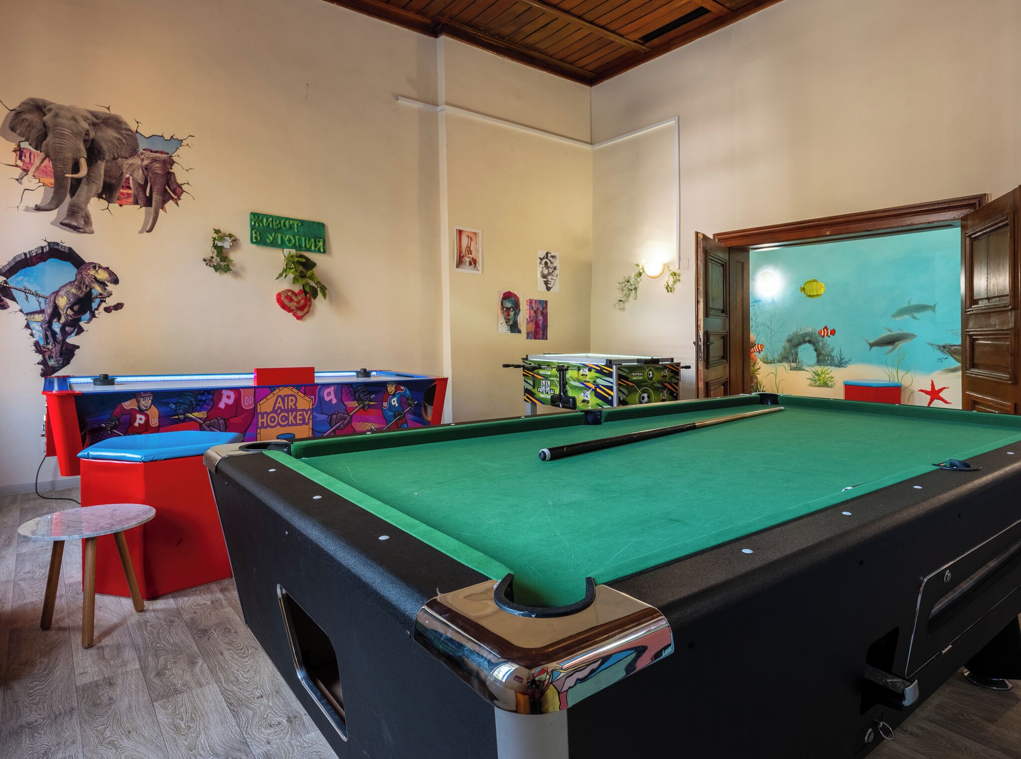 A pool table in a room with a wooden ceiling, a foosball table, and a wall with various decorations including a dinosaur mural and a sign with Cyrillic text.