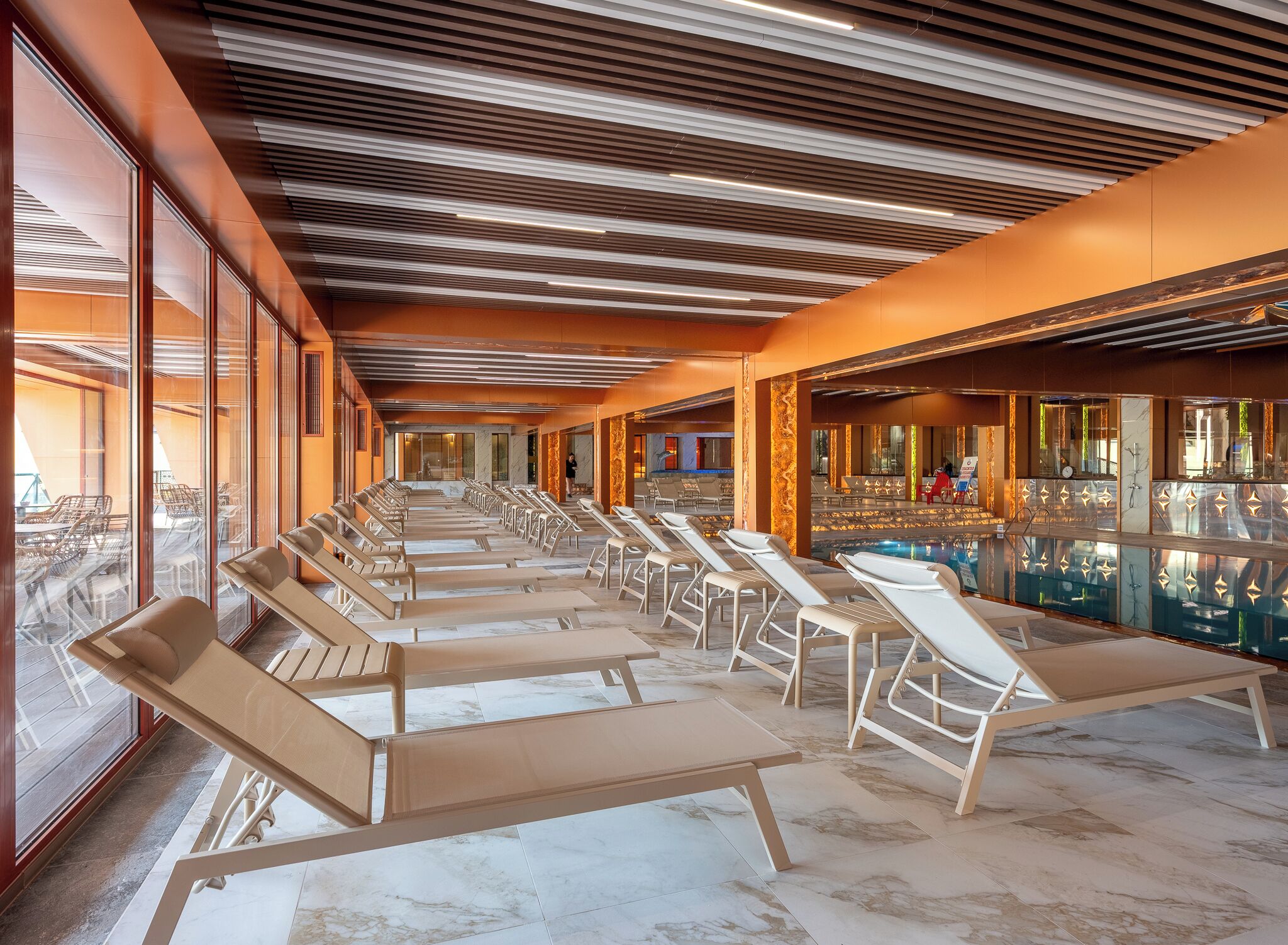 Zona d'una piscina interior il·luminada per la llum del sol, amb fileres de gandules i vista a la piscina a través d'un mur de vidre.