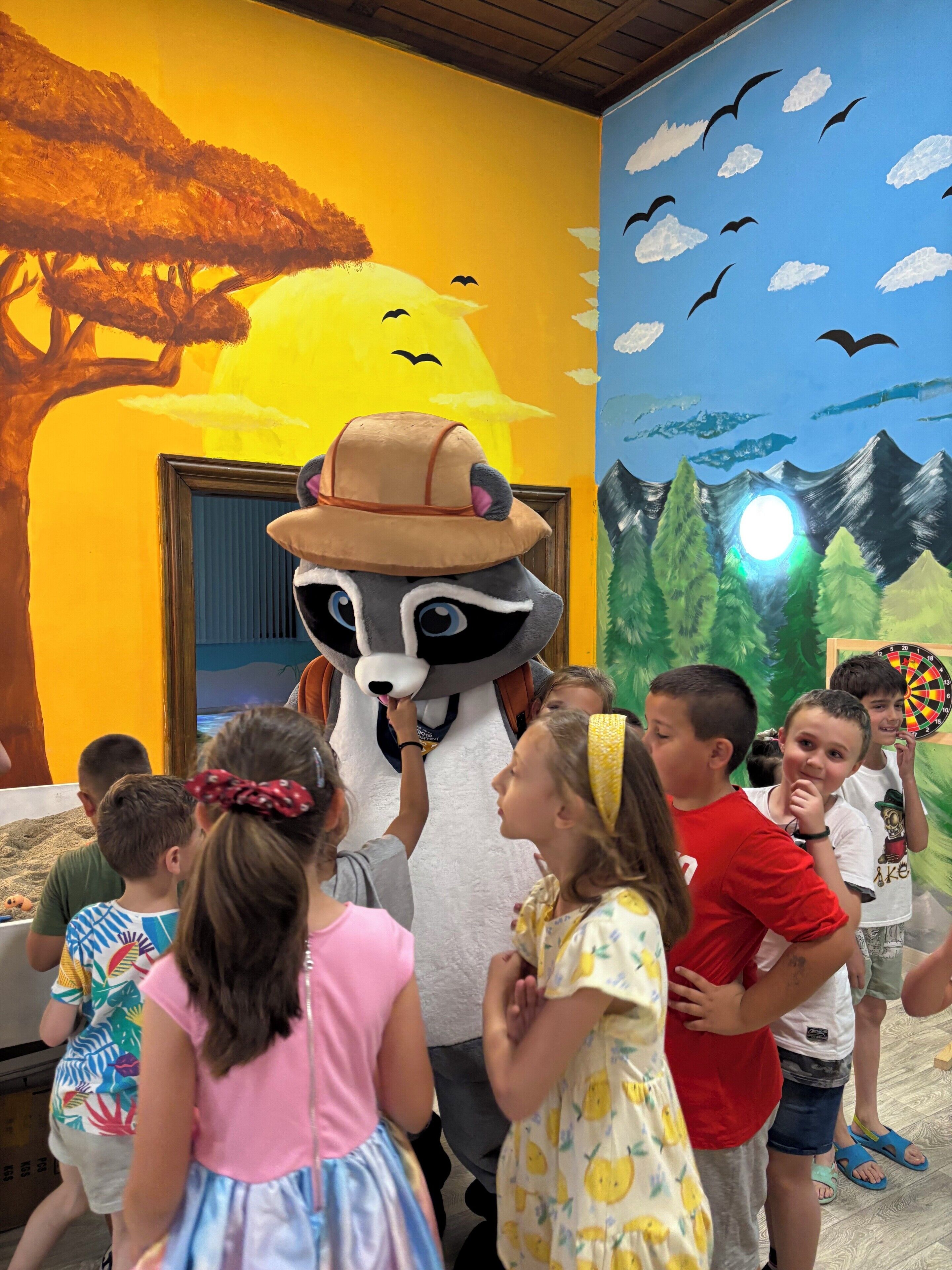 A room with a mural of a sunny landscape, a raccoon mascot, and children interacting.