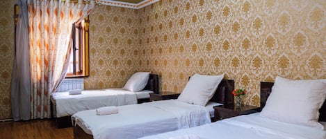 Habitación de hotel con tres camas, suelo de madera, papel tapiz estampado y un ventilador de techo.