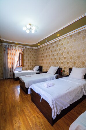 A hotel room with three beds, wooden flooring, patterned wallpaper, and a ceiling fan.