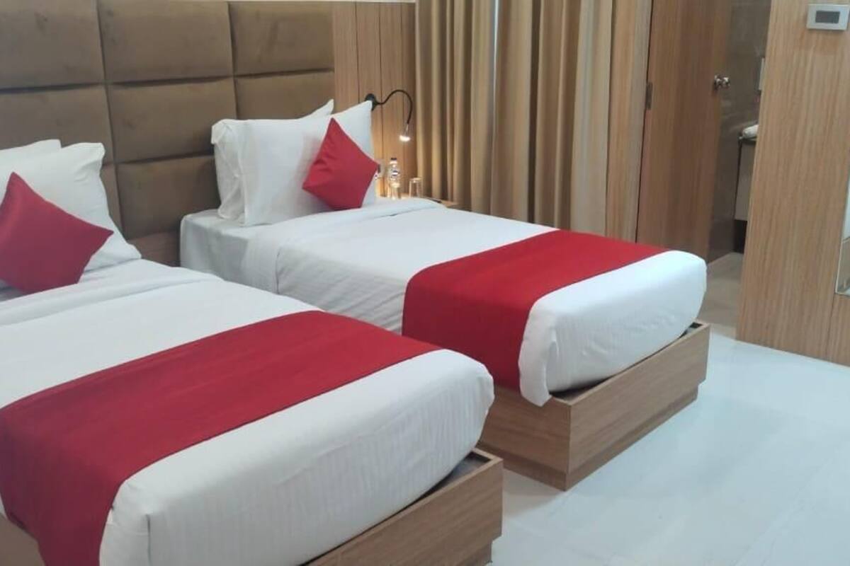 A hotel room with two beds, each with white bedding and red accents, a wooden headboard, a bedside table with a lamp, and a mirror on the wall.