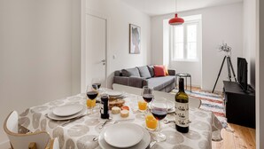 Ein Essbereich mit einem für zwei Personen gedeckten Tisch, einer Weinflasche und Gläsern Orangensaft. Im Hintergrund sind ein graues Sofa und eine rote Pendelleuchte zu sehen.
