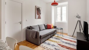 Ein modernes Wohnzimmer mit einem grauen Sofa, einem farbenfrohen Teppich, einem schwarzen Fernseher auf einem Stativ und einem gerahmten Kunstwerk an der Wand.