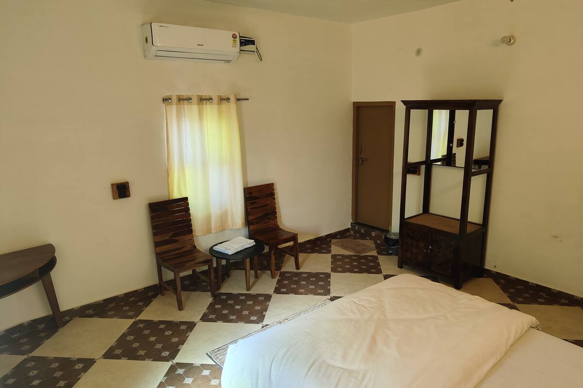A hotel room with a bed, two wooden chairs, a small table, an air conditioning unit, a mirror, and a door.