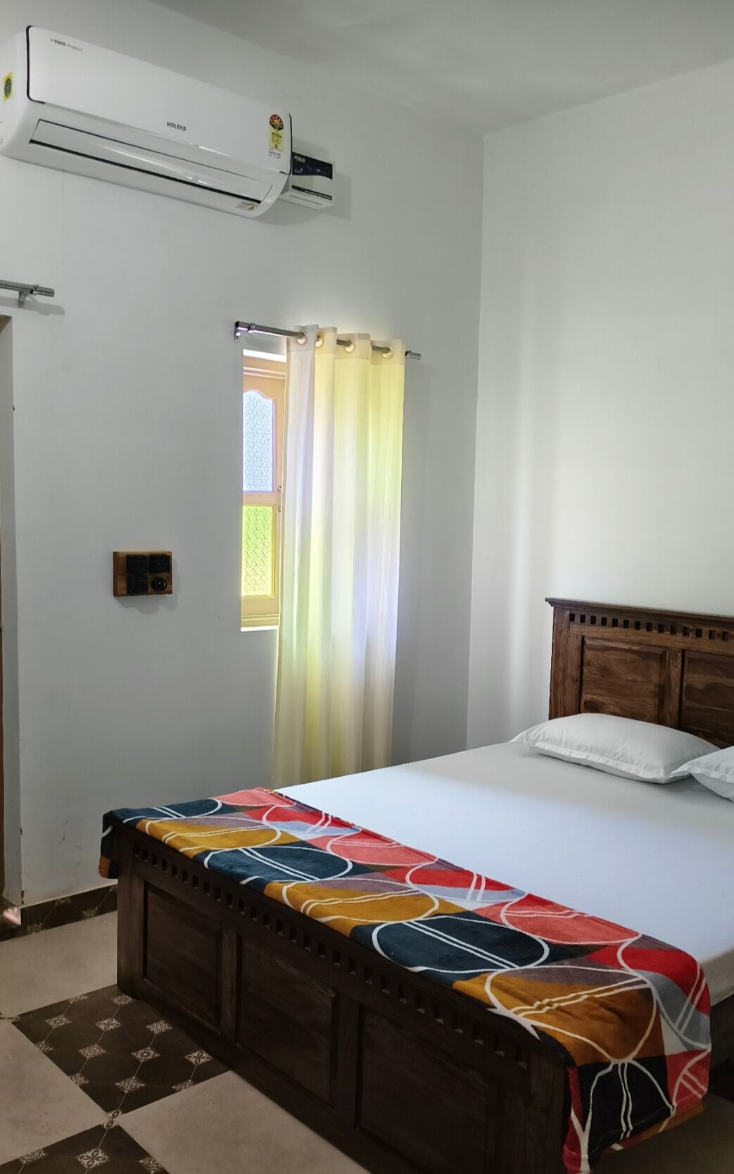 Een slaapkamer met een bed, een houten hoofdeinde, een raam met gordijnen, een airconditioning en een klein nachtkastje.
