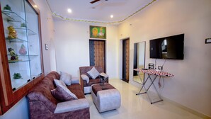 A living room with a brown leather sofa, a white ottoman, a glass cabinet with decorative items, a wall-mounted TV, and a ceiling fan. - SES LINGA COTTAGE (Palayankottai)