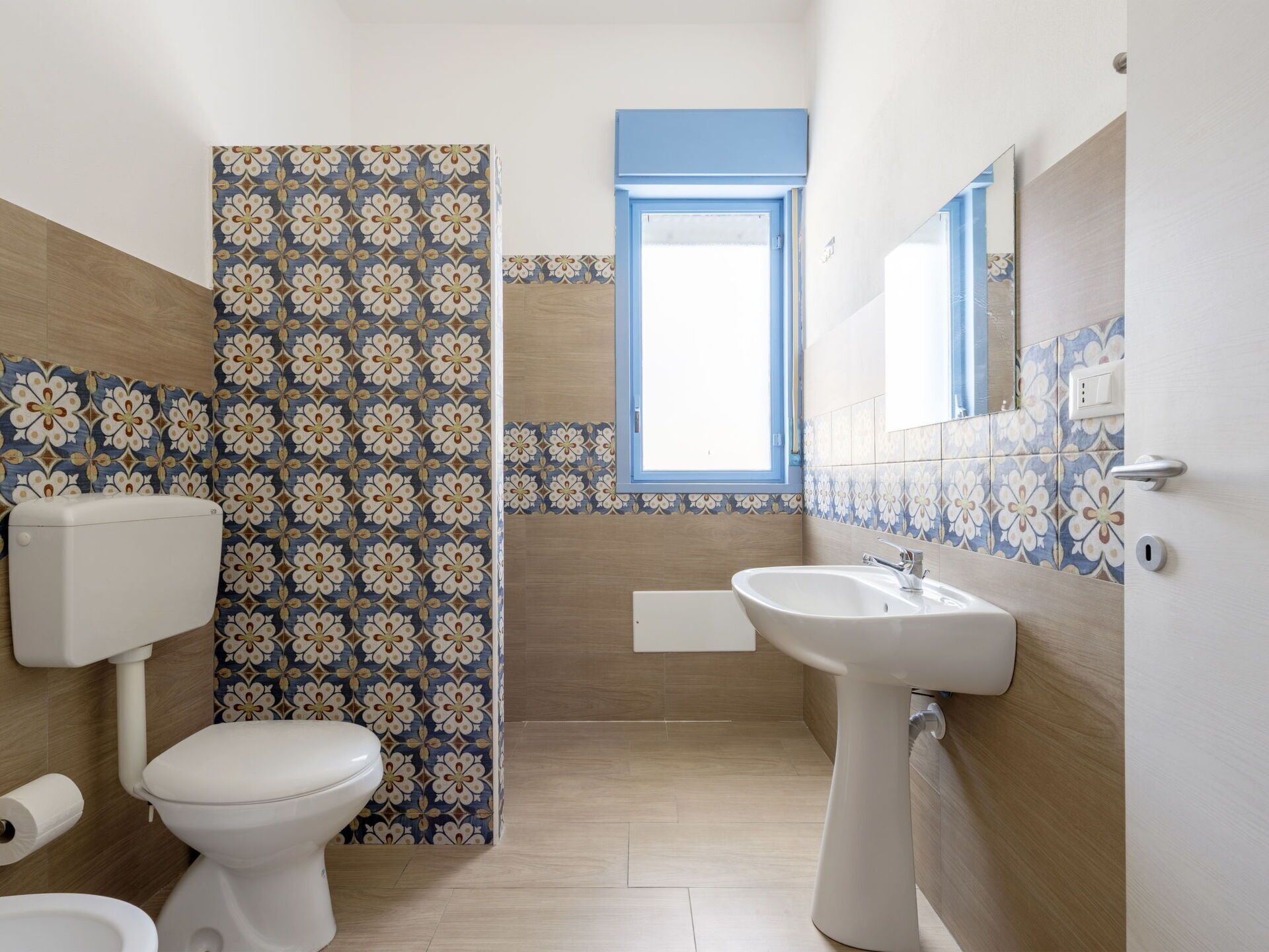 A bathroom with a toilet, bidet, and sink, featuring floral patterned tiles and a window.