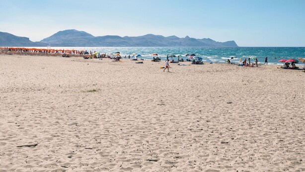 Een strand met mensen, parasols en bergen op de achtergrond.