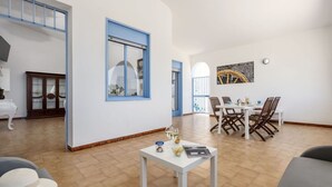 Een ruime woonkamer met een eettafel, stoelen, een witte salontafel en een groot raam met blauwe omlijsting.
