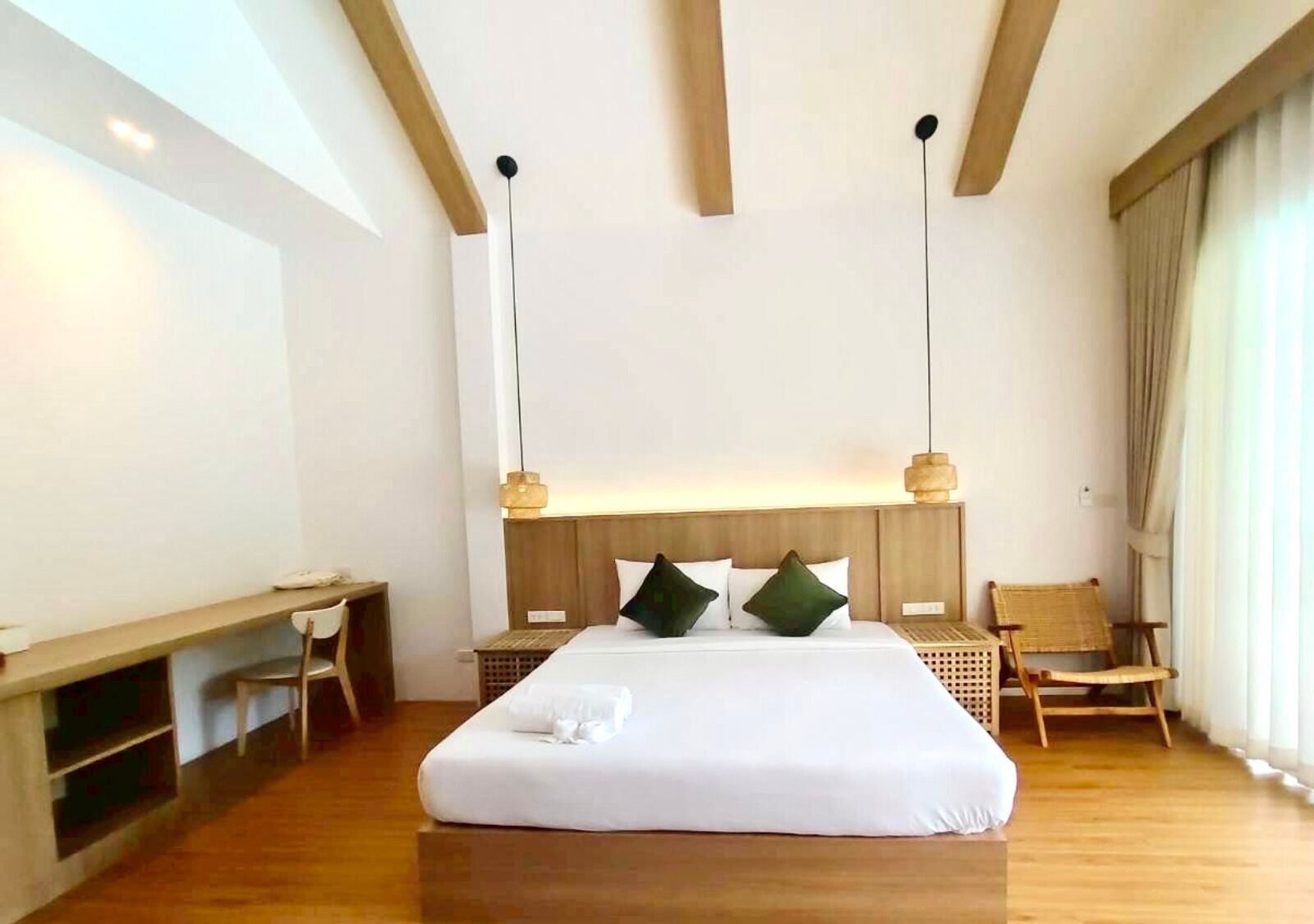 Ein modernes Hotelzimmer mit einem großen Bett, einem Holz-Kopfteil, zwei Nachttischen, einem Schreibtisch, einem Stuhl und einem großen Fenster mit Vorhängen.