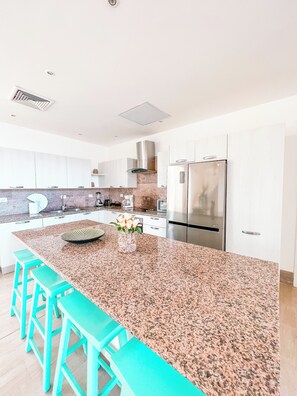 A modern kitchen with a large granite island, stainless steel appliances, and teal bar stools.