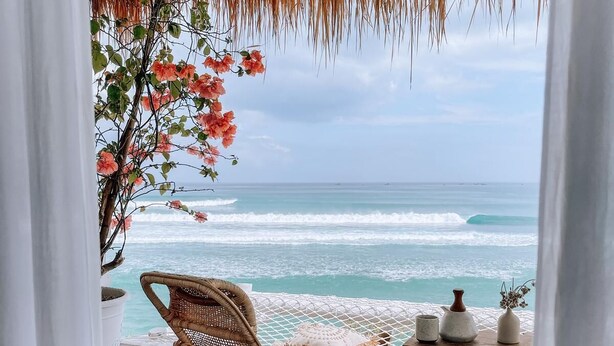 A wooden deck with a wicker chair and a small table with decorative items, overlooking a beach with waves and a clear sky. - Villa Deeksha tropicale ocean view Bingin (Bingin Kuta Selatan)