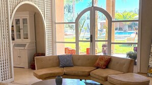 A living room with a beige sectional sofa, a round coffee table, and a view of a pool and garden through large windows. - Palais Bohemia (Zouhria)