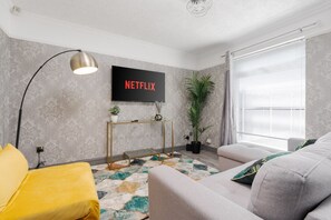 Un salón moderno con un sofá gris, un sillón amarillo, una mesa de centro de vidrio, una consola dorada, una planta grande en maceta y un televisor de pantalla plana montado en la pared.