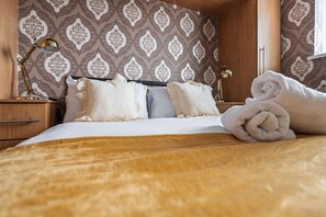 Una cama bien tendida con sábanas blancas y dos almohadas, una mesita de noche de madera con una lámpara, y un papel tapiz estampado.