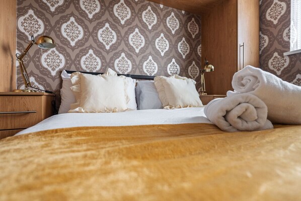 Ein ordentlich bezogenes Bett mit weißen Leinen und zwei Kissen, ein Nachttisch aus Holz mit einer Lampe sowie Wandpapier mit Muster.