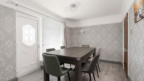 A dining room with a wooden table and four chairs, a clock on the wall, and patterned wallpaper. - Boutique 3bd Opposite Etihad & Coop - Free Parking (Greater Manchester)